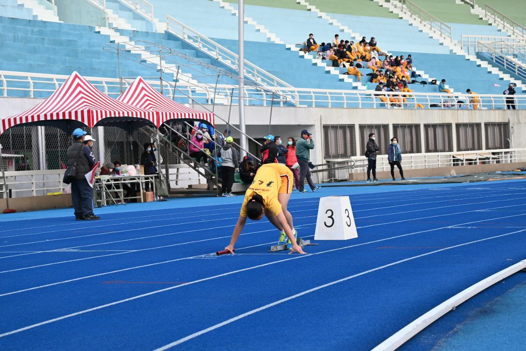 田徑隊選手在藍色PU跑道蹲踞起跑，手握接力棒準備衝刺